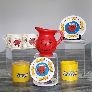 May include: A collection of vintage Kool-Aid kitchenware, including a red pitcher with a smiling face, plates, cups, and a yellow sugar container. The plates and cups feature the Kool-Aid Man logo.