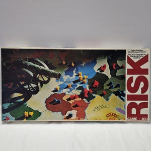 May include: A vintage Risk board game box. The box features a world map with territories and game pieces. A cannon is depicted on the left side. The word "RISK" is printed vertically in red, with "GAME JEU" below. The box is in good condition.