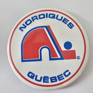 May include: A circular white button featuring a red and blue logo. The logo includes a red 'A' shape with blue detailing, a blue dot, and the words 'Nordiques' and 'Québec' in blue. A red ring borders the button.