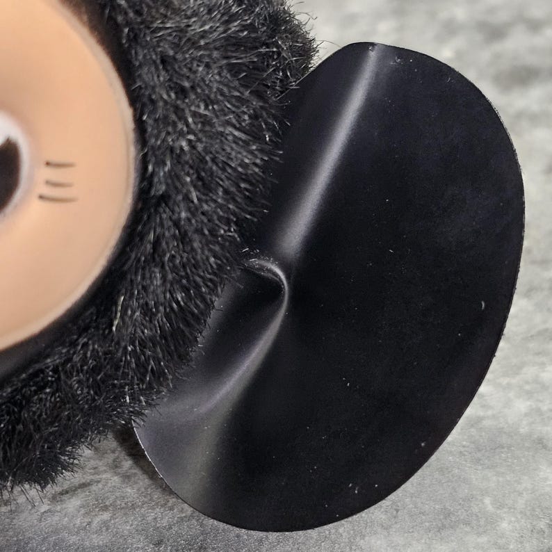 May include: Close-up of a black, rounded ear with a smooth, shiny surface. The ear is attached to a plush, black, furry head. The head has a tan face with three short, black lines.