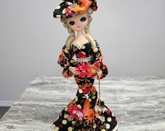 May include: A decorative doll wearing a floral print dress with a matching hat. The dress features a black background with red, orange, and yellow floral patterns and white polka dots. The doll has blonde hair and is holding a small fan.