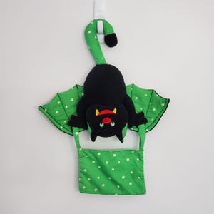 May include: A plush black bat toy with green wings and a green pouch, both featuring a small white star pattern. The bat has a red mouth, yellow fangs, and green eyes. It is suspended by a long green tail that curves upwards to form a hanging loop.