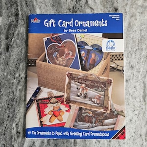 May include: A softcover instructional book titled Gift Card Ornaments by Bess Daniel, featuring a blue header and a cover display of painted tin ornaments including heart shapes, a snowman, and a gingerbread figure, with 17 projects included.