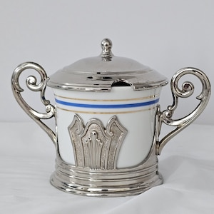 May include: A white porcelain sugar bowl with a silver lid and base. The bowl has two handles and a decorative silver design on the front. The rim features blue and gold stripes. The lid has a small knob and a slot for a spoon.
