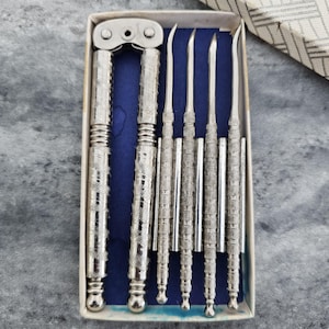 May include: A silver-toned metal nutcracker set featuring one hinged nutcracker tool and four slender, pointed picks. Each piece has a textured, ridged handle design. The set is arranged inside a rectangular box with a dark blue velvet lining.