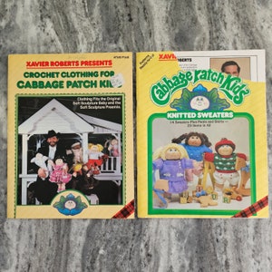 May include: Two vintage books on a gray surface. The book on the left is titled "Crochet Clothing for Cabbage Patch Kids" and features a photo of Xavier Roberts. The book on the right is titled "Cabbage Patch Kids Knitted Sweaters".