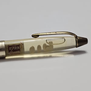 May include: A vintage clear pen with a gold-colored clip and tip. The pen's barrel contains several small, round, white tablets and a label that reads "Aspirin Genuine."