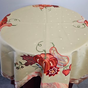 May include: A round beige tablecloth featuring a subtle white polka dot pattern and embroidered autumn motifs. The design includes orange pumpkins, gourds, red leaves, and flowers along the border, finished with a decorative marbled trim.
