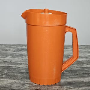 May include: A solid orange plastic jug featuring a cylindrical body, a sturdy side handle, and a moulded pouring spout. The top includes a circular lid with a small central knob for easy removal, and the base has a ribbed texture detail.