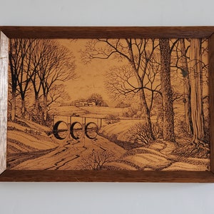May include: A framed landscape art piece featuring a detailed pen and ink drawing of a rural scene. The artwork depicts a bridge over a stream, trees, and fields, all rendered in shades of brown and black. The frame is made of dark wood.