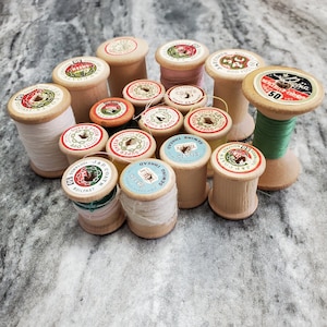 May include: A collection of vintage wooden spools of thread in various colors, including pink, green, and cream. The spools have colorful labels with text and designs, and some have thread still wound around them. A nostalgic image for sewing enthusiasts.