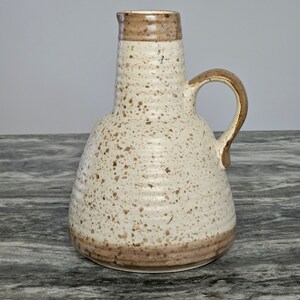 May include: A ceramic jug with a wide base and a narrow neck, featuring a cream-coloured body with brown speckles throughout. The rim and the curved handle are finished in a solid brown glaze, and the exterior surface has horizontal ridges.