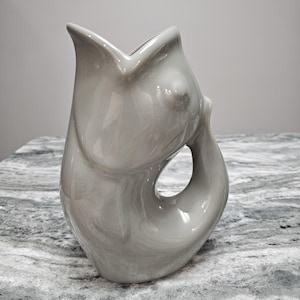 May include: A gray ceramic vase with a unique, organic shape. The vase has a large opening and a curved handle-like design with a circular cutout. The surface has a glossy finish with subtle marbling, adding visual interest to the decorative piece.