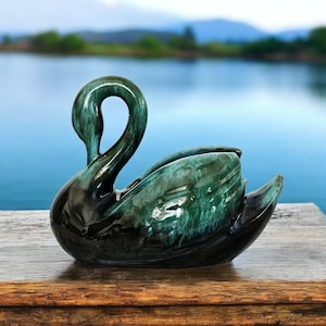 May include: A ceramic swan figurine featuring a glossy finish with a deep emerald green and black mottled glaze. The swan is depicted with its neck gracefully curved downward, showcasing a smooth, rounded body and detailed wing textures.