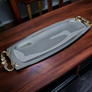 May include: A rectangular silver serving tray with gold-coloured handles. The tray is highly reflective, with a polished surface. The handles have an ornate, curved design. The tray is placed on a dark wooden surface.