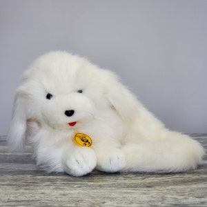 May include: A fluffy white plush dog toy with black eyes and a red tongue. The toy has long, floppy ears and a yellow tag with a panda bear logo. The dog is lying down on a wooden surface.