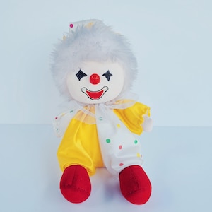 May include: A plush clown doll with a white face, black diamond-shaped eyes, and a red nose and mouth. The doll wears a yellow and white outfit with red shoes and a fluffy white headpiece. The outfit has colorful polka dots.
