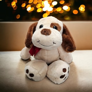 May include: A soft toy dog with a cream body, brown ears, and a brown patch above one eye. It has a red bow and a smiling face. The background features blurred golden lights.