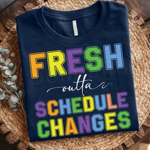May include: Navy blue t-shirt with the text "FRESH outta SCHEDULE CHANGES" in colorful block letters. The shirt is displayed on a woven mat with a wooden background. The design includes the text "d mal DESIGNS".