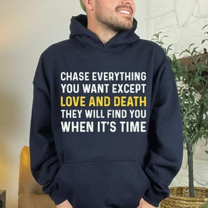 以下が含まれることがあります： 「CHASE EVERYTHING YOU WANT EXCEPT LOVE AND DEATH THEY WILL FIND YOU WHEN IT'S TIME」という白い文字がプリントされた、ネイビーブルーのパーカー。 ドローストリングフードとフロントポケットが付いています。