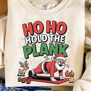 Op de afbeelding: Crèmekleurige sweatshirt met een kerstthema. De afbeelding toont de kerstman die een plankoefening doet, met de tekst "HO HO HOLD THE PLANK" in rood en groen. Een rendier, gewichten en ornamenten zijn ook inbegrepen.