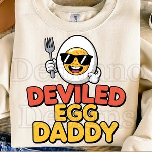 May include: Cream-colored sweatshirt with the text "DEVILED EGG DADDY" in red and yellow. The graphic features a cartoon deviled egg wearing sunglasses and holding a fork. The bottom of the image reads "300DPI/ SVG + PNG/DEGITAL FILE".