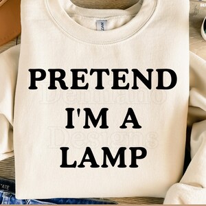 Könnte beinhalten: Cremefarbenes Sweatshirt mit dem Text "PRETEND I'M A LAMP" in fetten schwarzen Buchstaben. Das Sweatshirt hat einen Rundhalsausschnitt und lange Ärmel. Der untere Teil des Bildes zeigt den Text "300DPI / SVG+PNG / DEGITAL FILE".