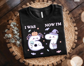 May include: Black t-shirt with the text "I WAS 6, NOW I'M 7" and Halloween-themed graphics. The design includes a ghost, a pumpkin, a witch's hat, and candy. The shirt is on a woven mat.