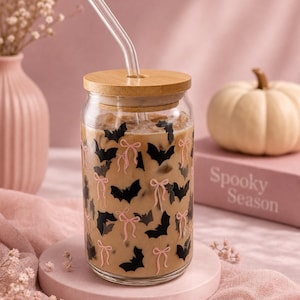 May include: A clear glass tumbler with a bamboo lid and a glass straw, filled with an iced beverage. The exterior of the glass features a repeating pattern of black bats and light pink bows printed across the surface of the cylindrical container.
