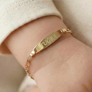 May include: A gold-colored bracelet with a rectangular nameplate and a chain link band. The nameplate is engraved with the name "TUĞBA". The bracelet is worn on a wrist, with a cream-colored sleeve in the background.