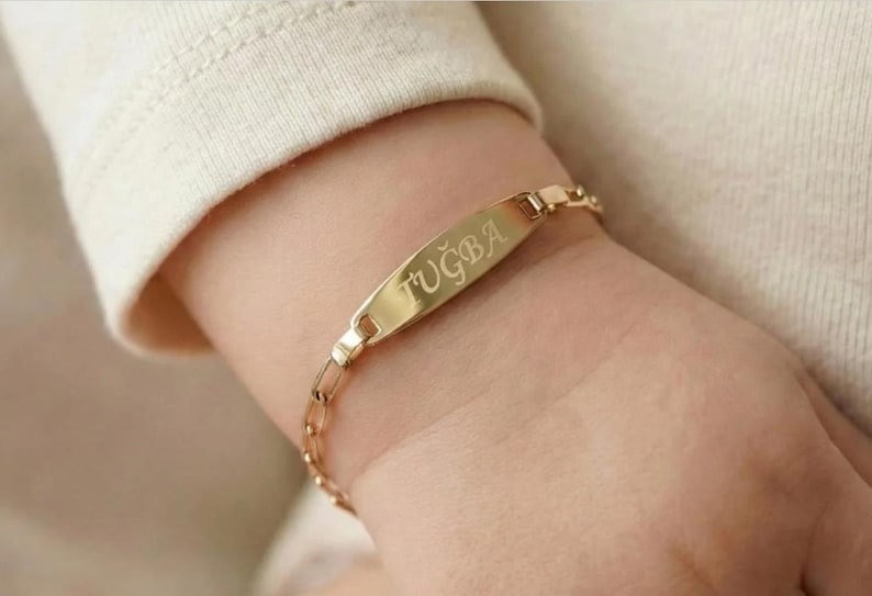 May include: A gold-colored bracelet with a rectangular nameplate and a chain link band. The nameplate is engraved with the name "TUĞBA". The bracelet is worn on a wrist, with a cream-colored sleeve in the background.