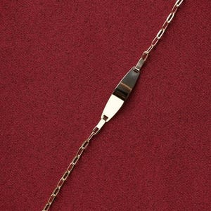May include: A gold-colored ID bracelet with a rectangular plate and a chain link design. The plate is slightly curved and has a polished finish. The bracelet is displayed on a red background.