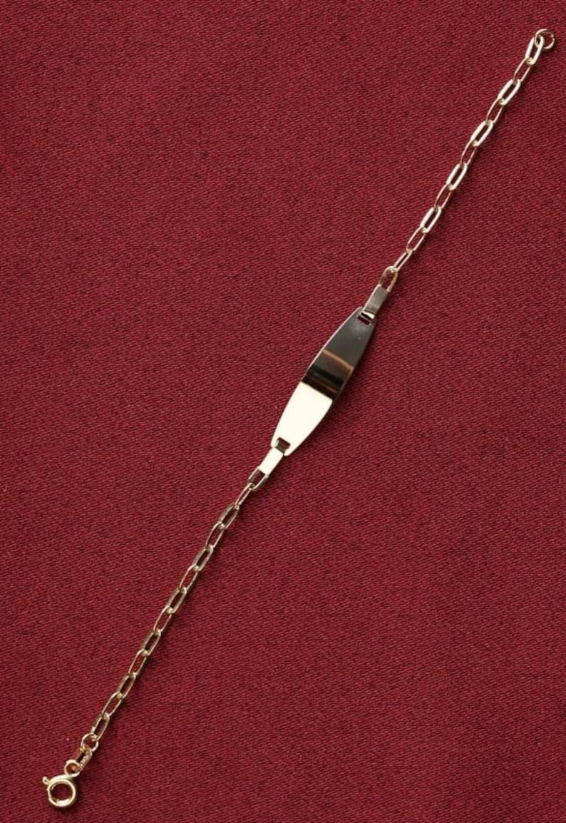 May include: A gold-colored ID bracelet with a rectangular plate and a chain link design. The plate is slightly curved and has a polished finish. The bracelet is displayed on a red background.