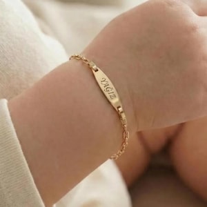 May include: A gold-colored bracelet with the name "YAGIZ" engraved on a rectangular plate. The bracelet is attached to a delicate chain. The bracelet is on a wrist.