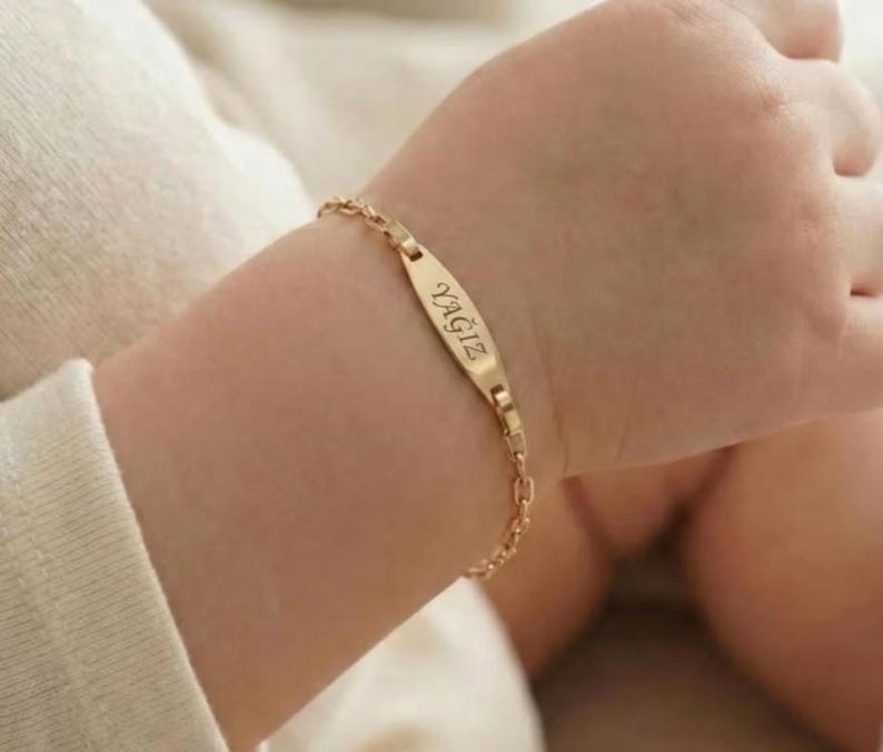 May include: A gold-colored bracelet with the name "YAGIZ" engraved on a rectangular plate. The bracelet is attached to a delicate chain. The bracelet is on a wrist.