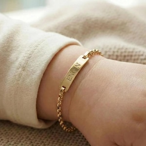 May include: A gold-colored bracelet with the name "ENGIN" engraved on a rectangular plate. The bracelet features a delicate chain design and is worn on a wrist.