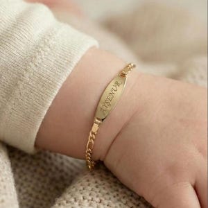 May include: A gold-colored baby bracelet with the name "AYŞENUR" engraved on a rectangular plate. The bracelet has a delicate chain design and is shown on a baby's wrist.