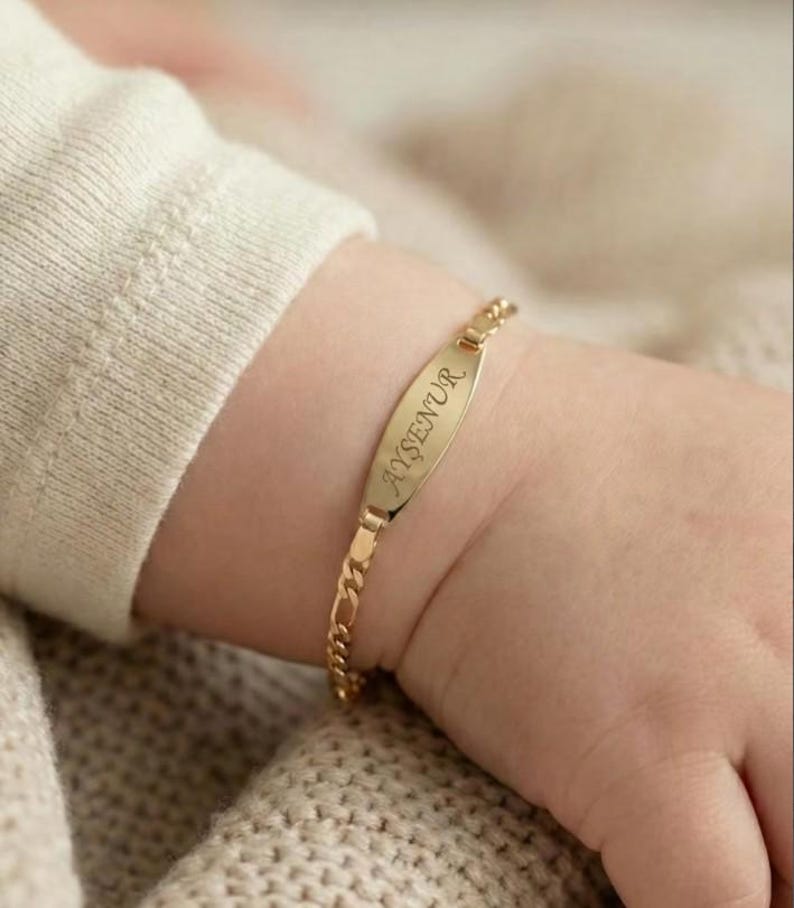 May include: A gold-colored baby bracelet with the name "AYŞENUR" engraved on a rectangular plate. The bracelet has a delicate chain design and is shown on a baby's wrist.