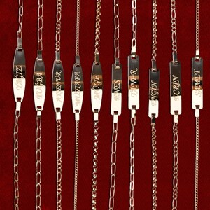 May include: Gold-colored name bracelets with various chain styles. Each bracelet features a rectangular plate with a name engraved on it. The bracelets are displayed against a red background. The image includes the product codes BLK1120 to BLK1111.