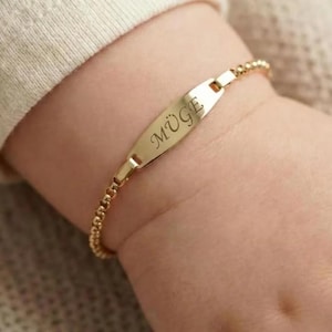 May include: A gold-colored bracelet with the name "M&Uuml;GE" engraved on a rectangular plate. The bracelet is on a baby's wrist. The bracelet is made of small, round beads.