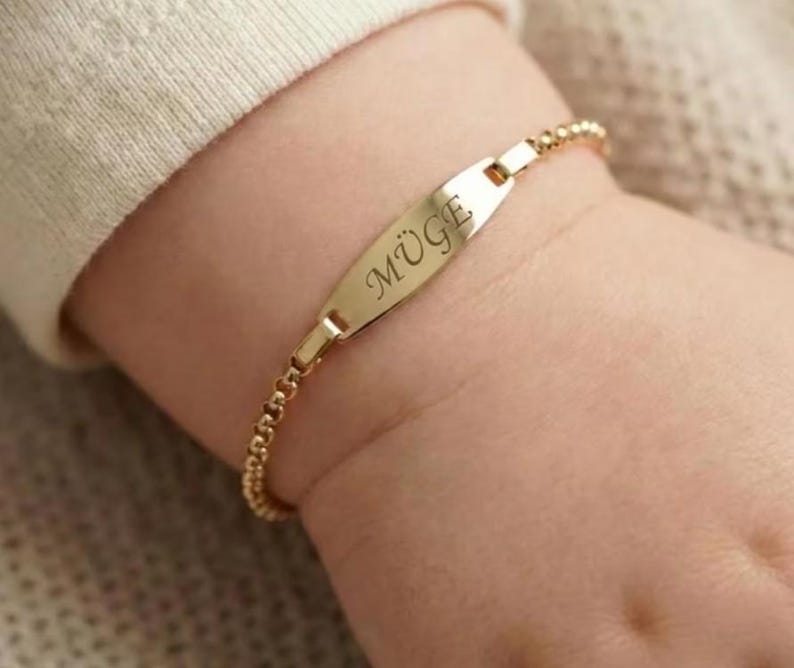 May include: A gold-colored bracelet with the name "M&Uuml;GE" engraved on a rectangular plate. The bracelet is on a baby's wrist. The bracelet is made of small, round beads.