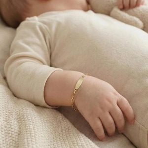 May include: A gold baby ID bracelet with a rectangular nameplate and a delicate chain. The bracelet is on a baby's wrist, lying on a cream-colored blanket. The baby is wearing a cream-colored onesie.