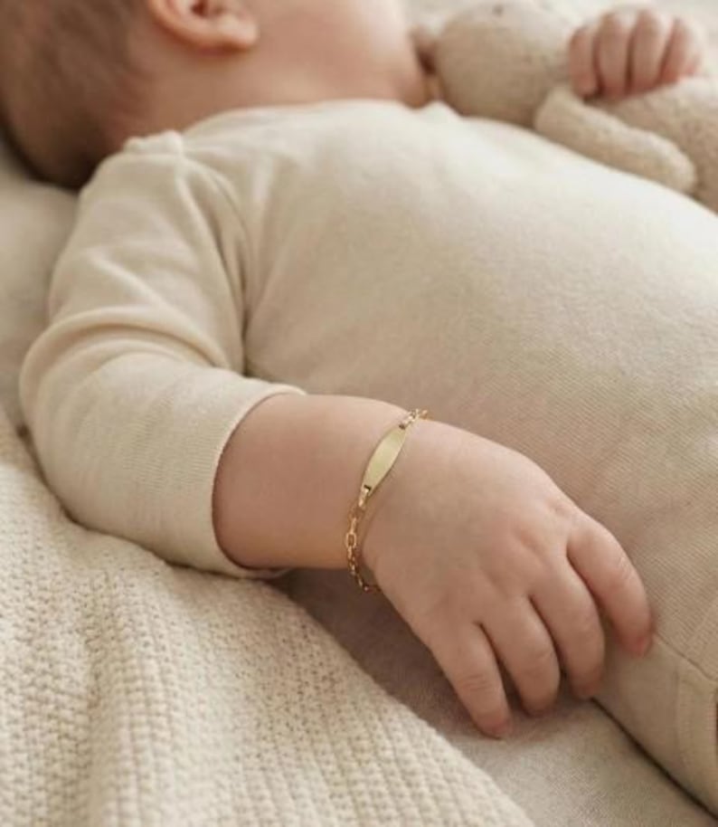 May include: A gold baby ID bracelet with a rectangular nameplate and a delicate chain. The bracelet is on a baby's wrist, lying on a cream-colored blanket. The baby is wearing a cream-colored onesie.