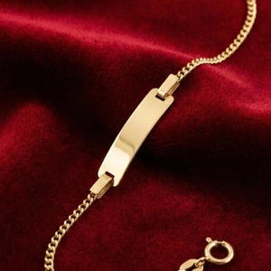 May include: A gold-colored ID bracelet with a rectangular plate and a curb chain. The bracelet is displayed on a red velvet surface. The clasp is a small, round spring ring.