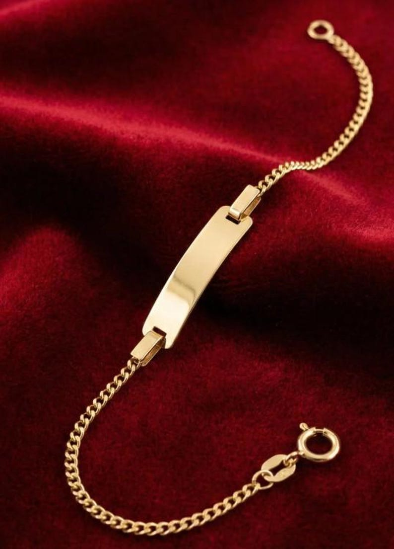 May include: A gold-colored ID bracelet with a rectangular plate and a curb chain. The bracelet is displayed on a red velvet surface. The clasp is a small, round spring ring.