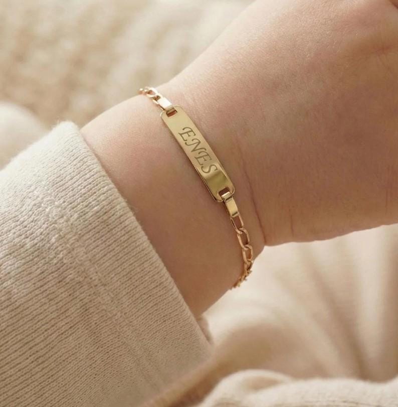 May include: A gold-colored bracelet with the name "ENES" engraved on a rectangular plate. The bracelet is on a child's wrist, and the chain links are visible. The background is a soft, neutral color.