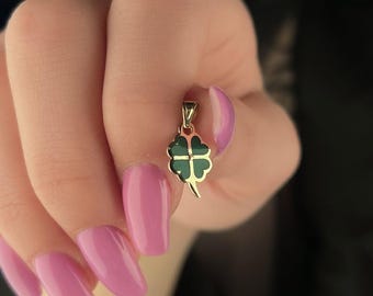 May include: Gold-colored pendant with a four-leaf clover design. The clover has a green enamel filling. The pendant is suspended from a small loop. The background is dark.