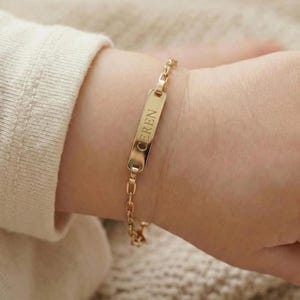 May include: A gold-colored bracelet with a rectangular nameplate engraved with the name "EREN". The bracelet is attached to a delicate chain and is worn on a wrist.