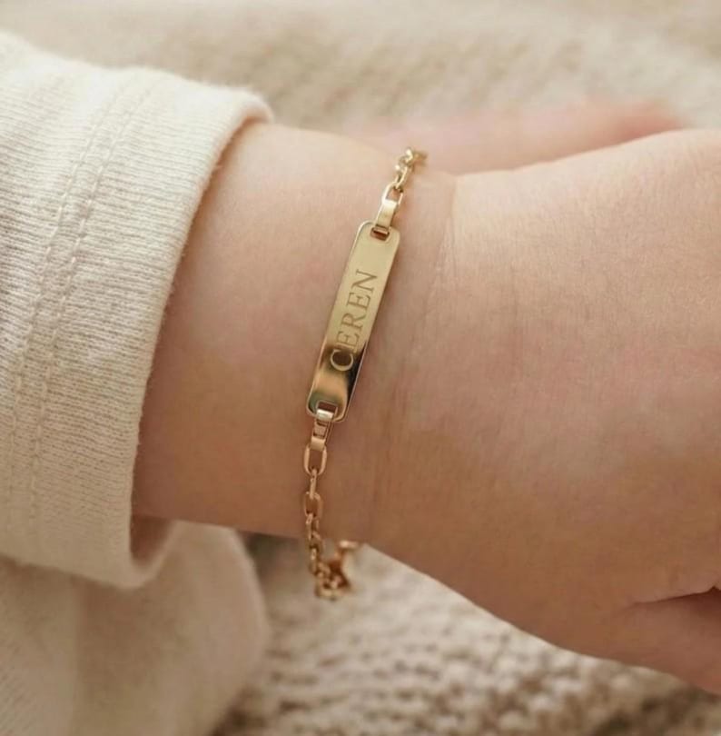 May include: A gold-colored bracelet with a rectangular nameplate engraved with the name "EREN". The bracelet is attached to a delicate chain and is worn on a wrist.