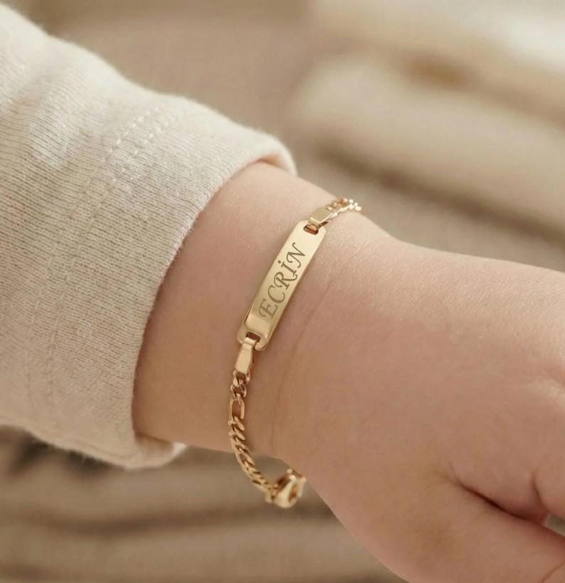 May include: A gold-colored bracelet with the name "ECRIN" engraved on a rectangular plate. The bracelet is on a child's wrist. The chain has a delicate, linked design.